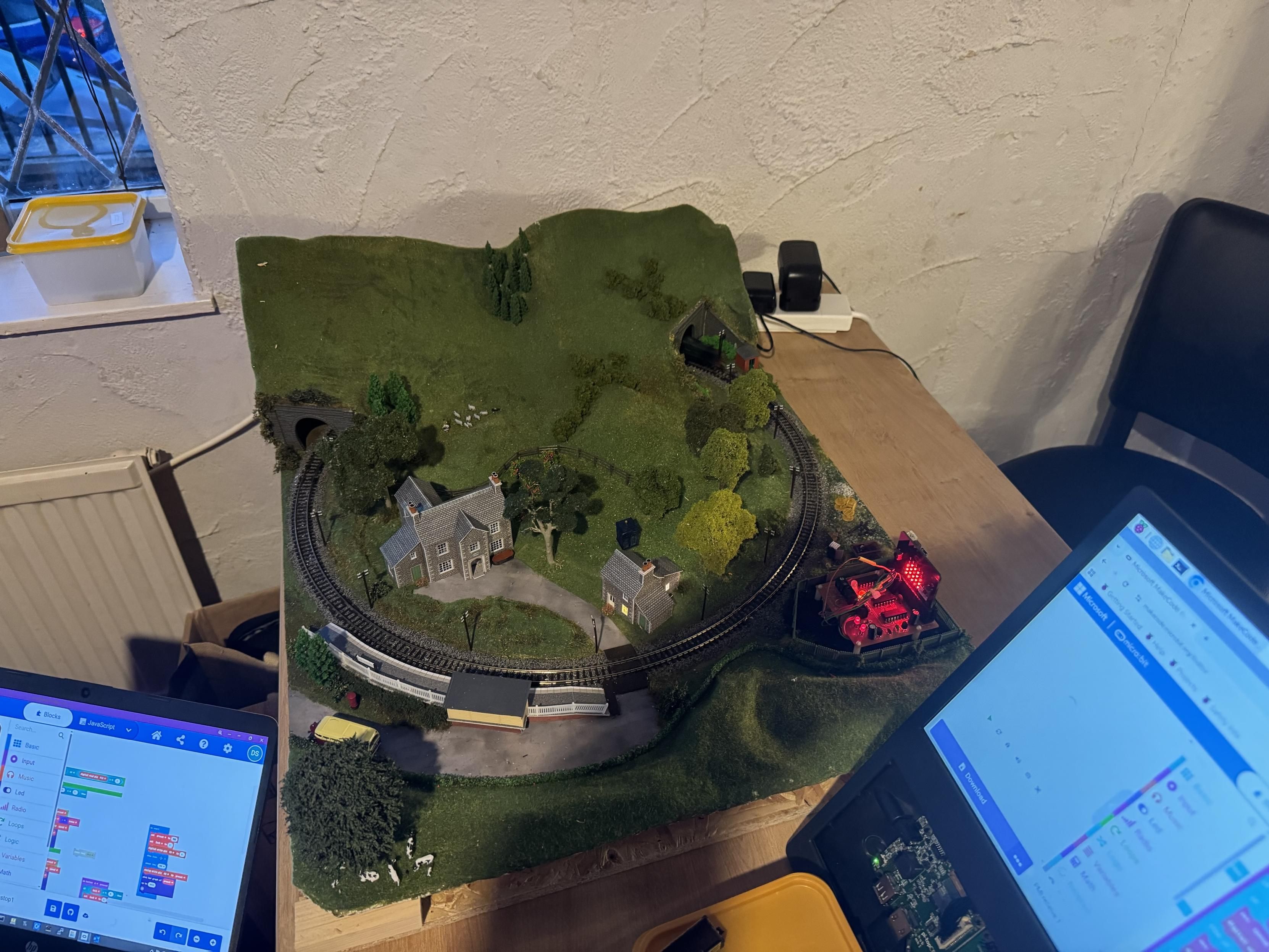 A tabletop model railway scene featuring a curved track, a small village with houses and trees, and a red-lit structure. Nearby, a laptop displays programming blocks, indicating ongoing digital setup or control for the model.
