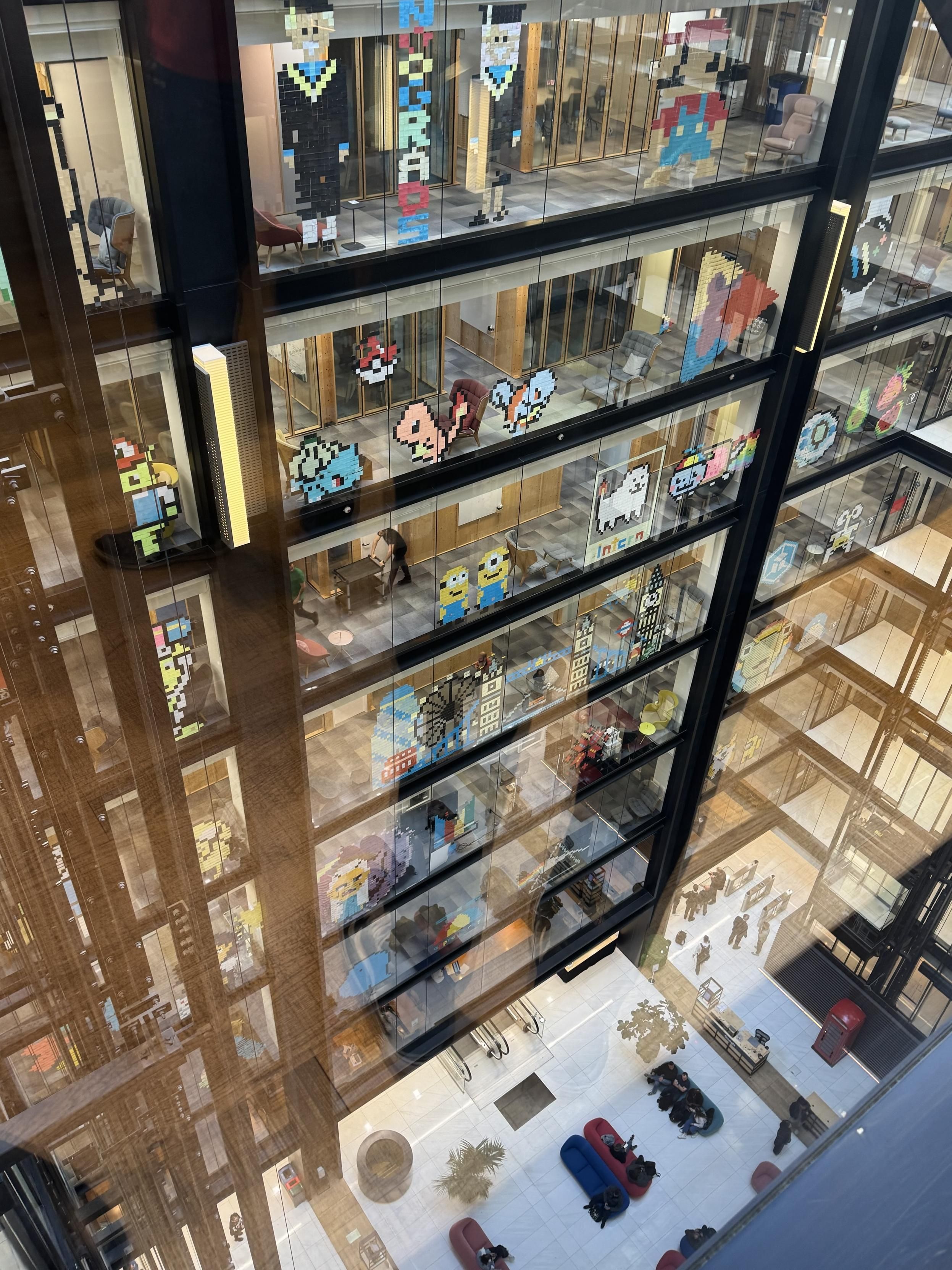 An interior view shows a multi-level building with large glass walls decorated with pixel art characters and designs. The floor below features seating areas, people, and decorative plants, creating a vibrant, creative atmosphere.