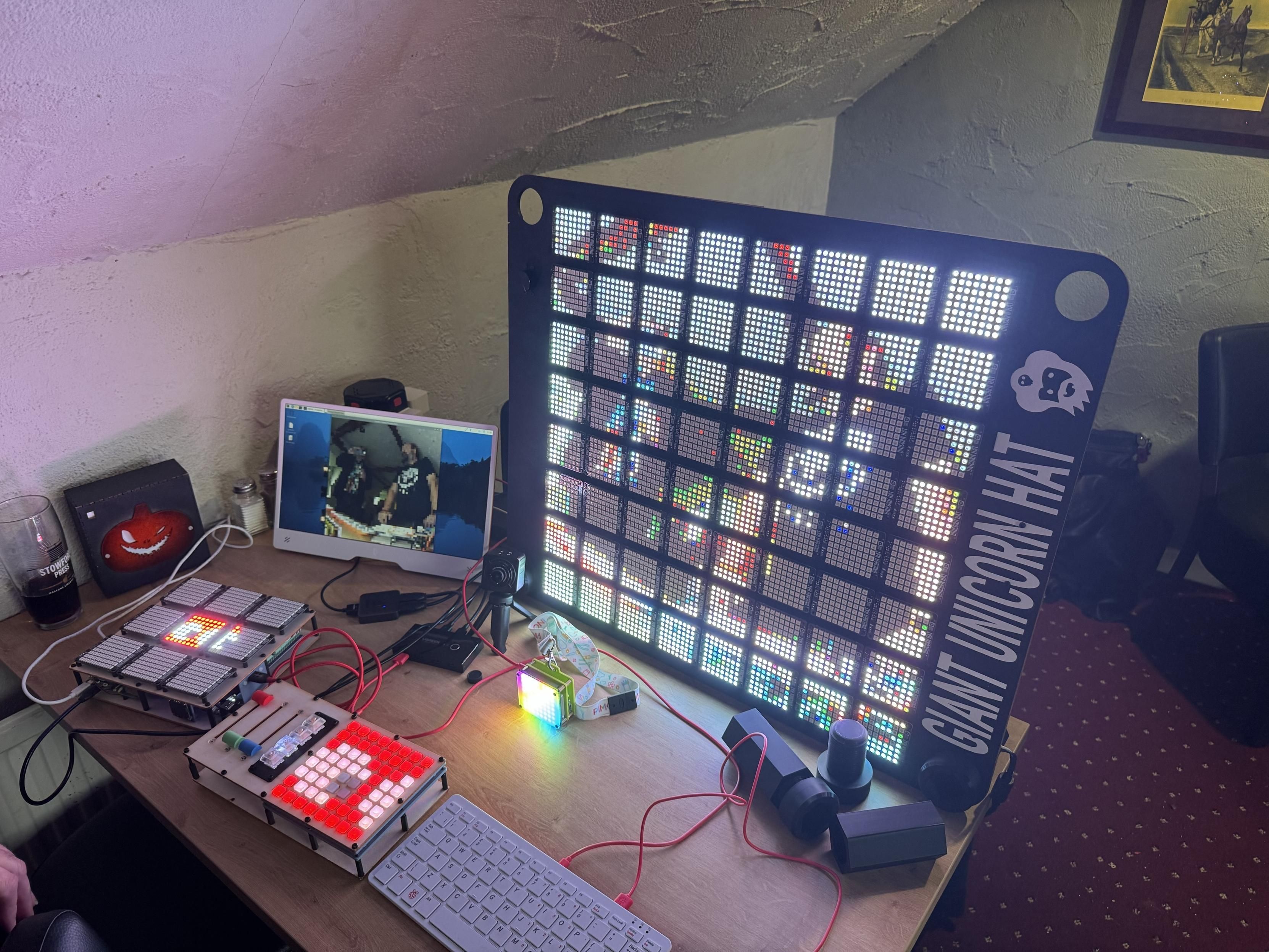 A workspace featuring a large LED display labeled "Giant Unicorn Hat," with colorful lights. There is a smaller LED panel below, both connected by wires. An adjacent monitor displays a scene with two individuals.